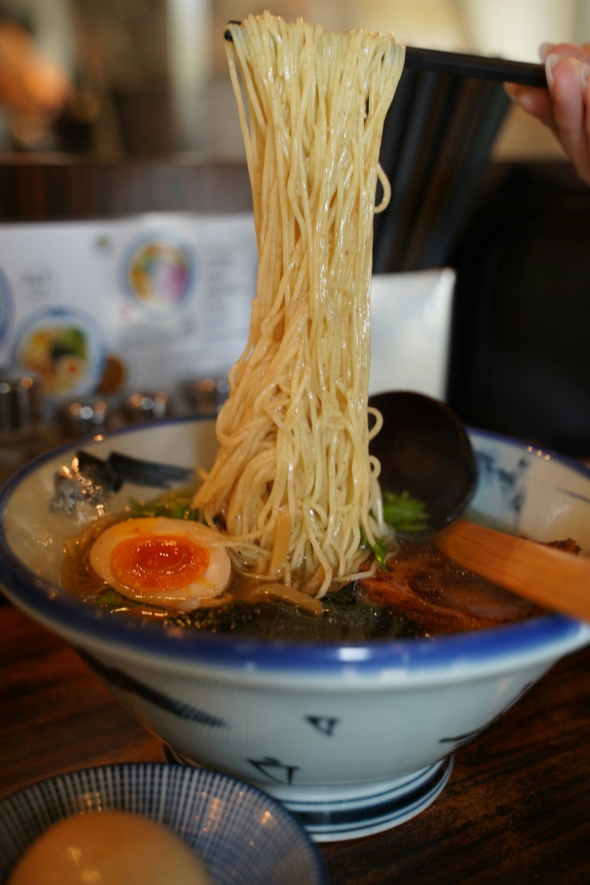 A bowl of ramen with chopsticks lifting noodles and a soft-boiled egg on a wooden table