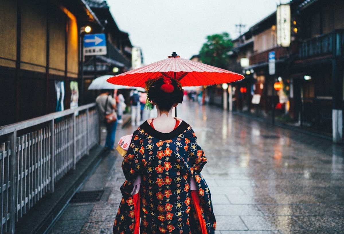 Woman in patterned kimono holding a red oil paper umbrella walking down a rain-wet Gion street, Kyoto Japan