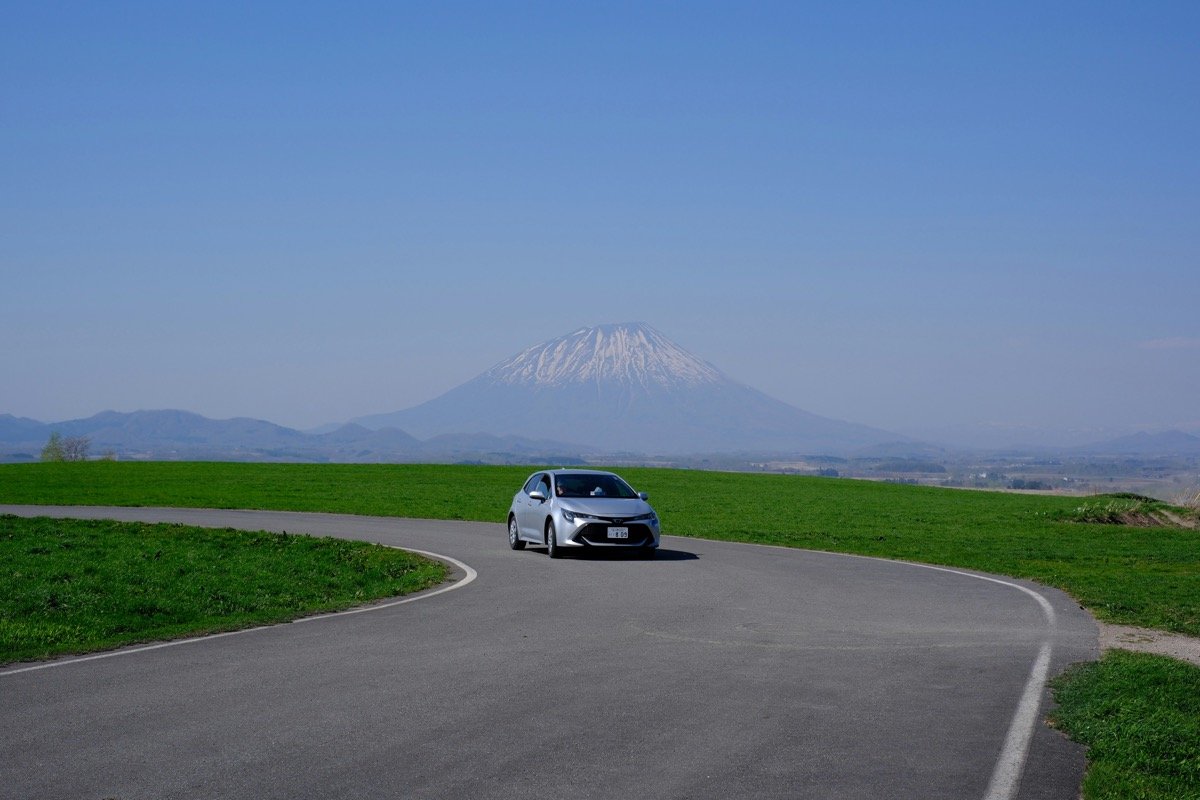 Silver car on a winding road in Hokkaido with a snow-capped volcano in the background