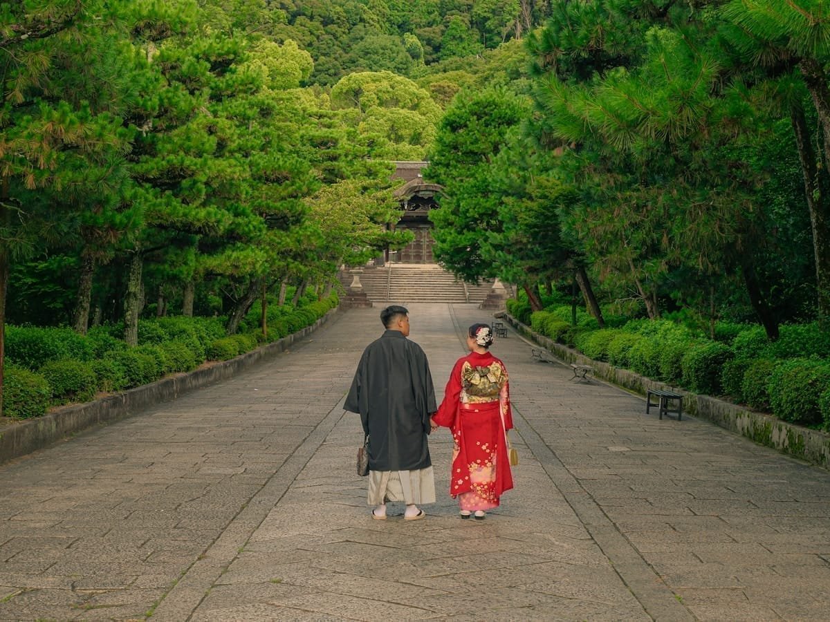 Couple in traditional kimonos walking hand in hand up a stone path toward a temple, surrounded by green trees, Japan